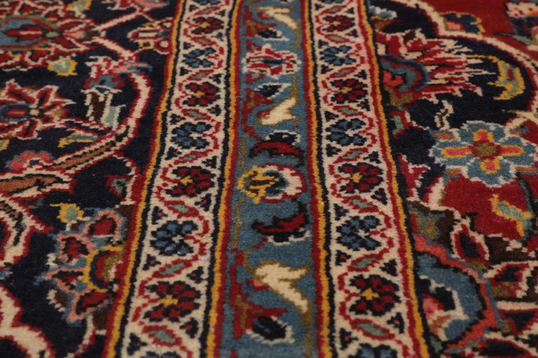 Een handgeknoopt Keshan-tapijt uit het Midden-Oosten met een rood veld en donkerblauwe randen.