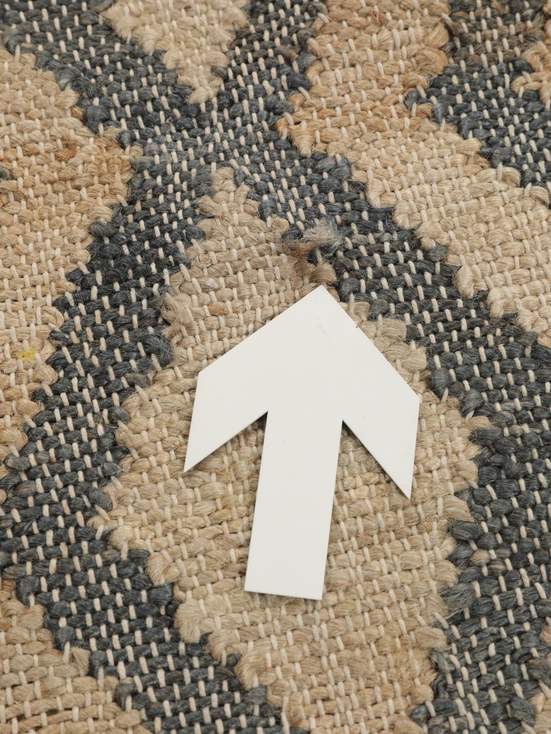 Um tapete retangular de juta tecido à mão, proveniente da Índia, com um padrão de losangos em bege e cinzento-escuro.