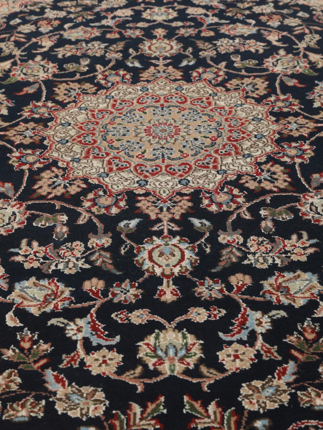 Un tapis rectangulaire Nain Indo présentant un champ bleu foncé avec un médaillon floral central et des bordures complexes, noué à la main en Inde.