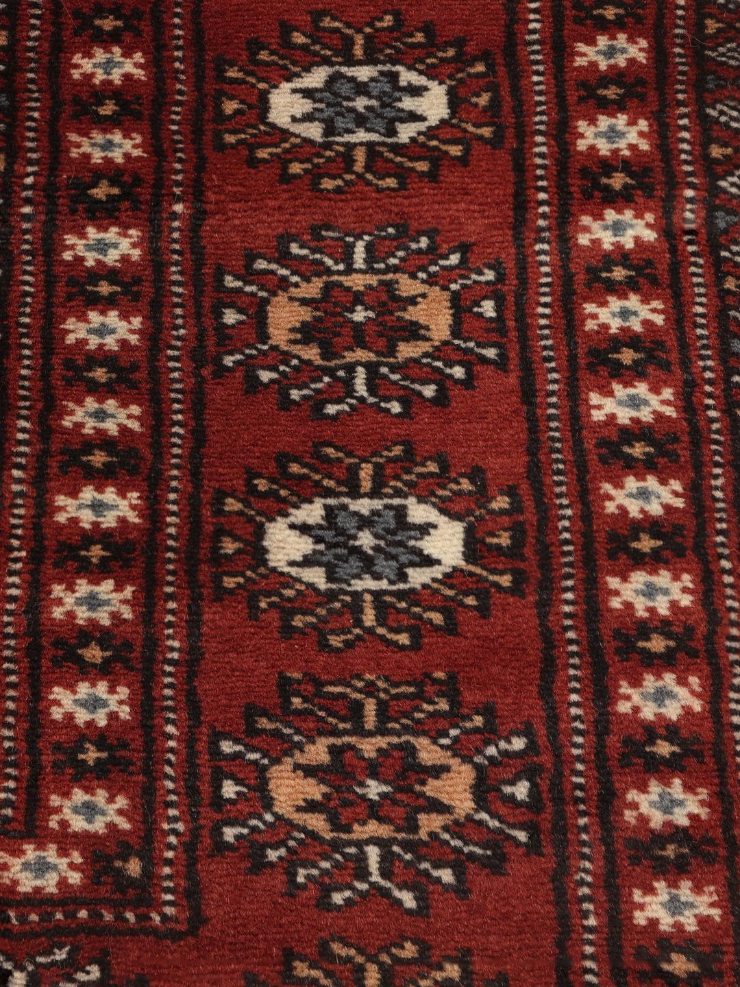 Un tapis Pakistan Bokhara noué à la main avec un champ rouge profond et des motifs géométriques répétitifs.