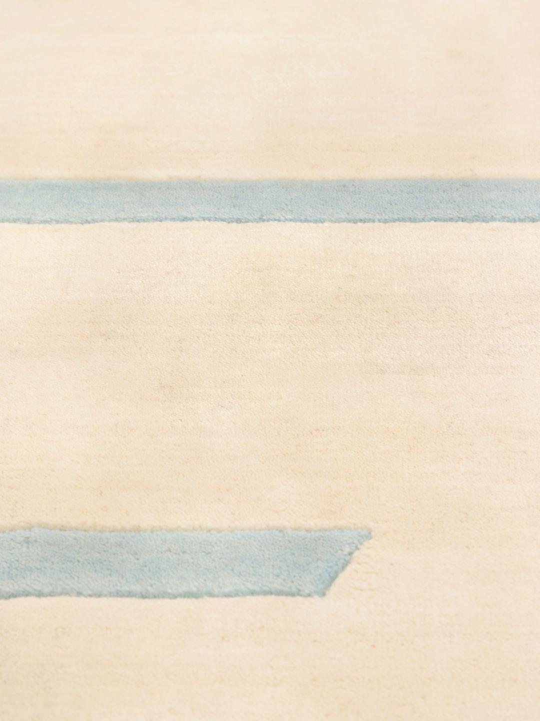 Et rektangulært håndlavet uldtæppe fra Indien med en beige base og lyseblå striber.