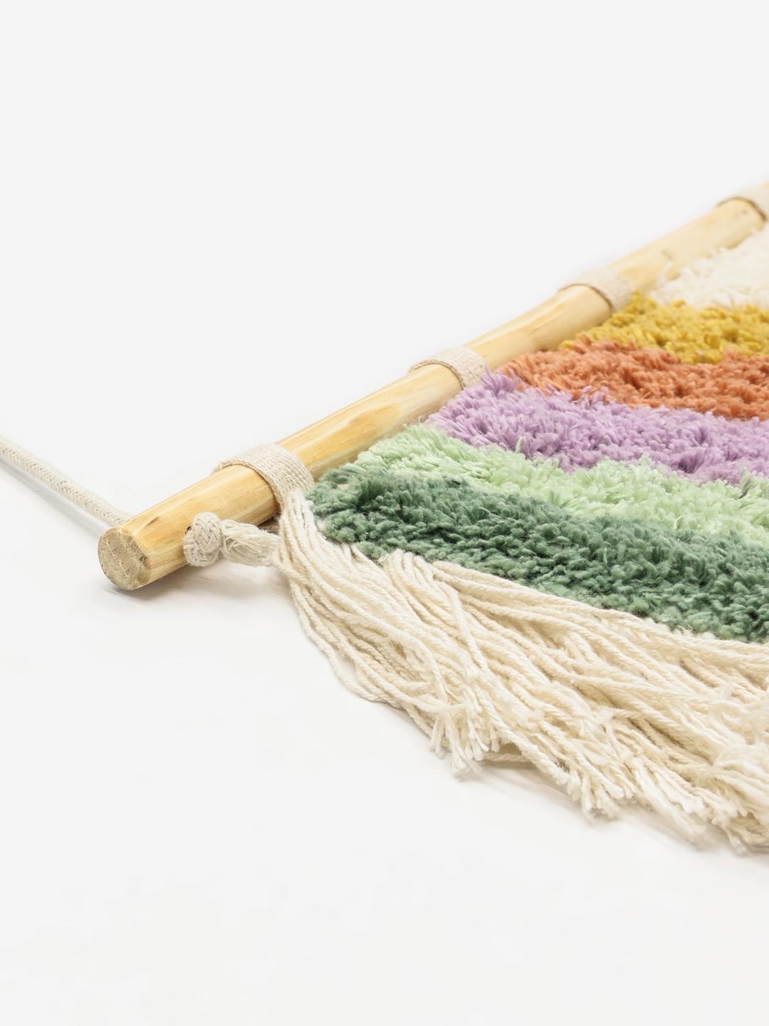 Een kleurrijke, halfronde regenboog wanddecoratie gemaakt van katoen met lange franjes die aan een houten roede hangt.