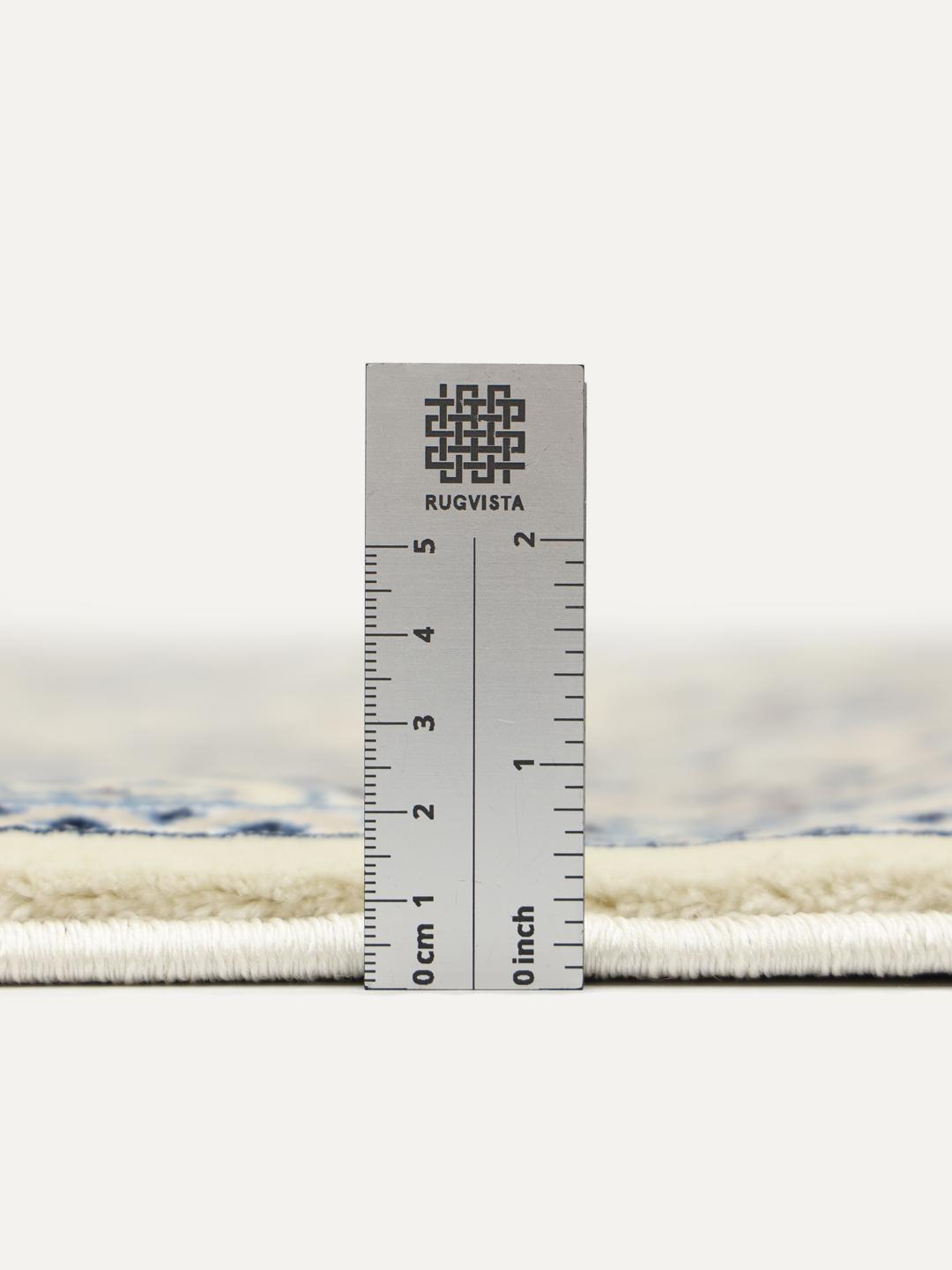 Rundt tæppe i beige og lyseblå med detaljerede blomstermotiver og frynsekant.