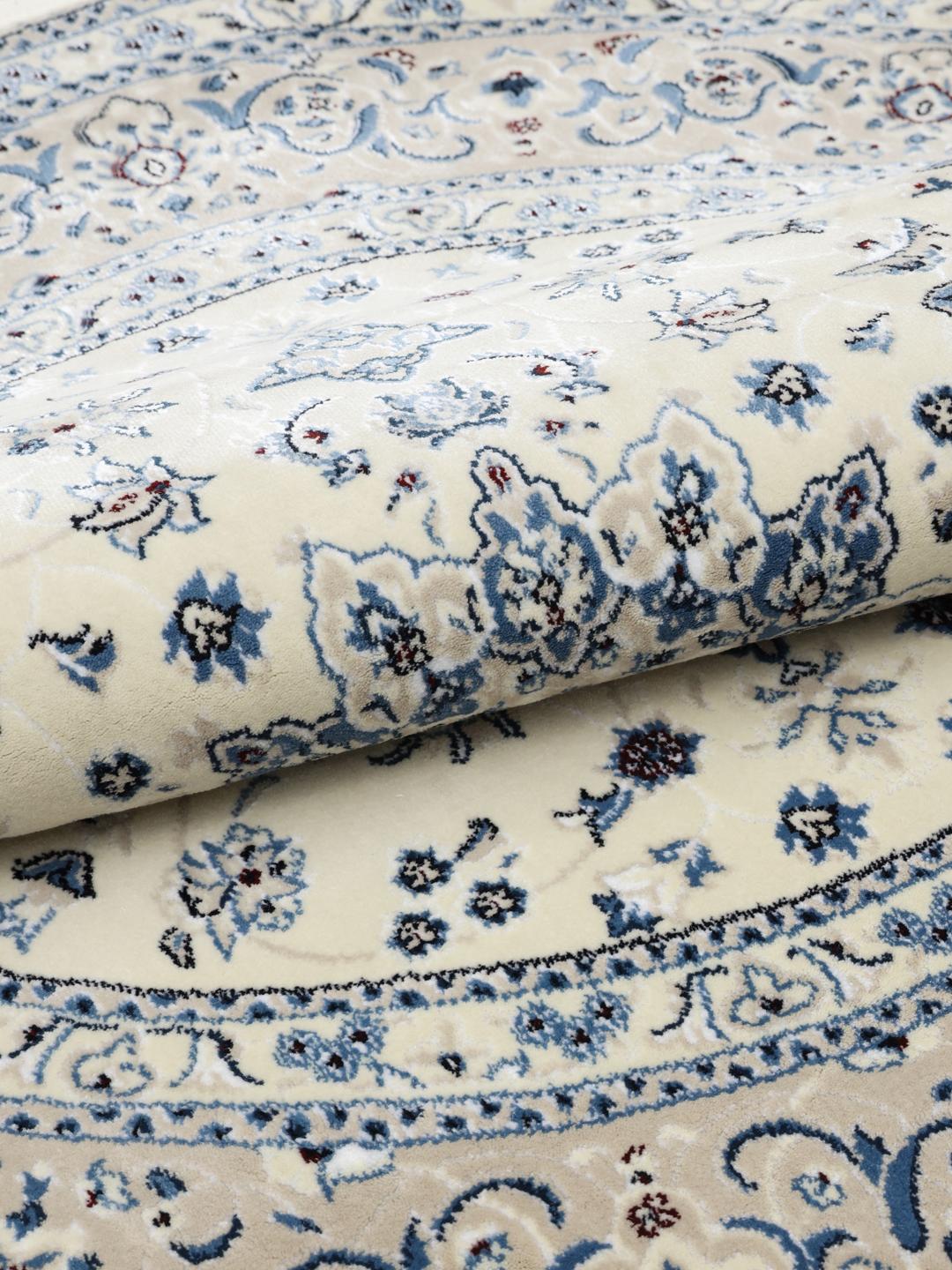 Rundt tæppe i beige og lyseblå med detaljerede blomstermotiver og frynsekant.