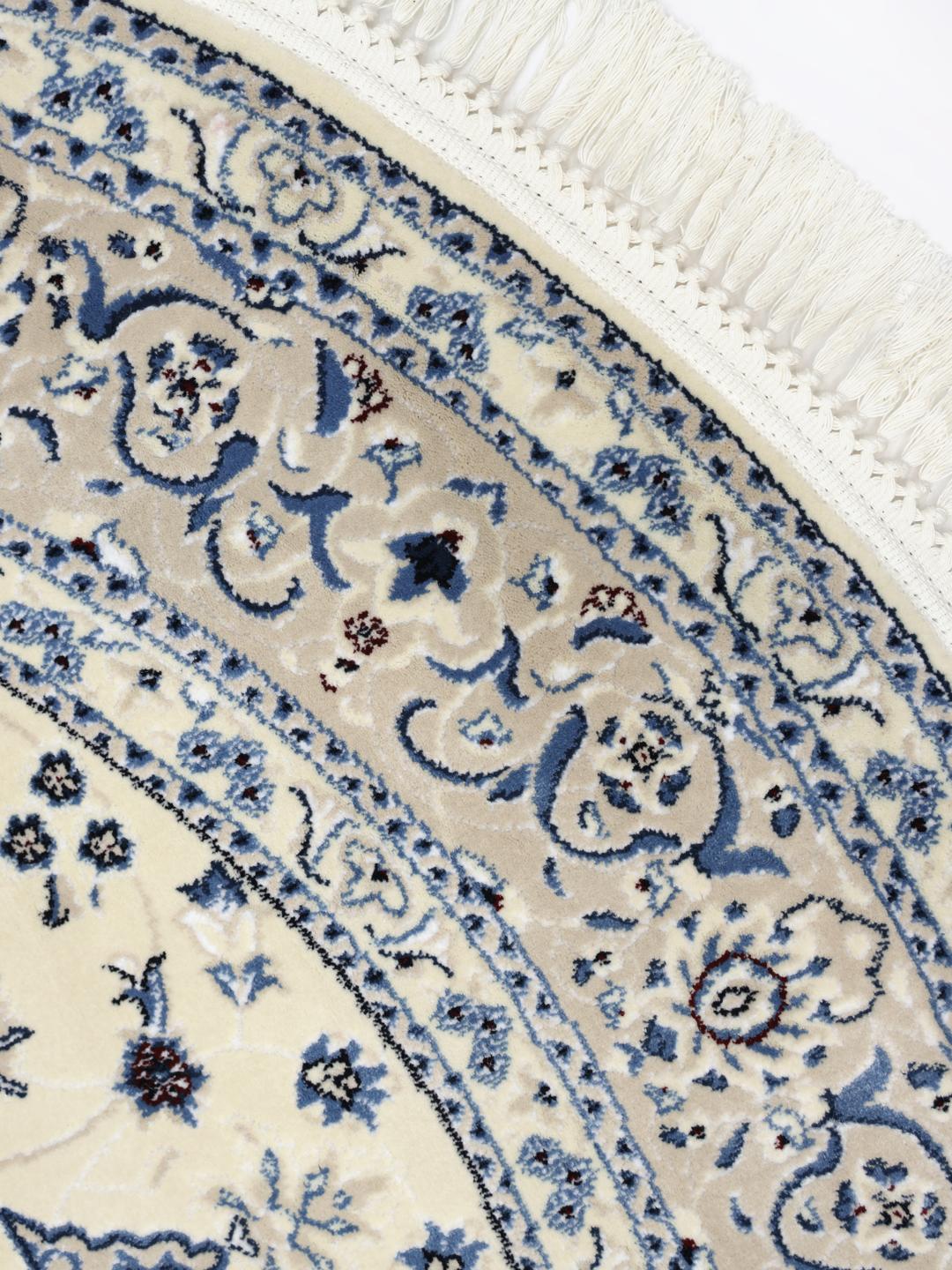 Rundt tæppe i beige og lyseblå med detaljerede blomstermotiver og frynsekant.