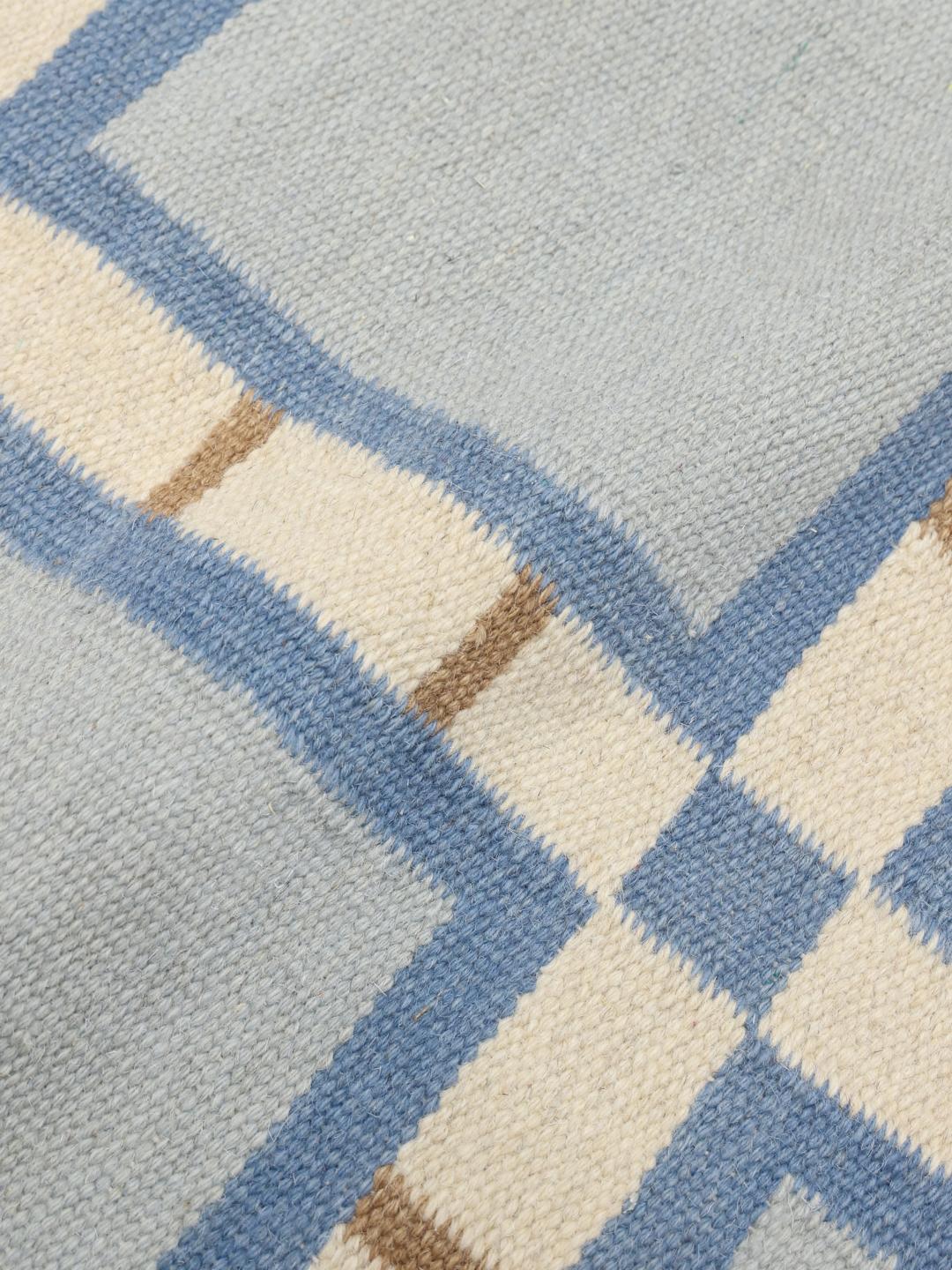 Et rektangulært uldtæppe med et geometrisk gittermønster i blå, beige og hvide nuancer.