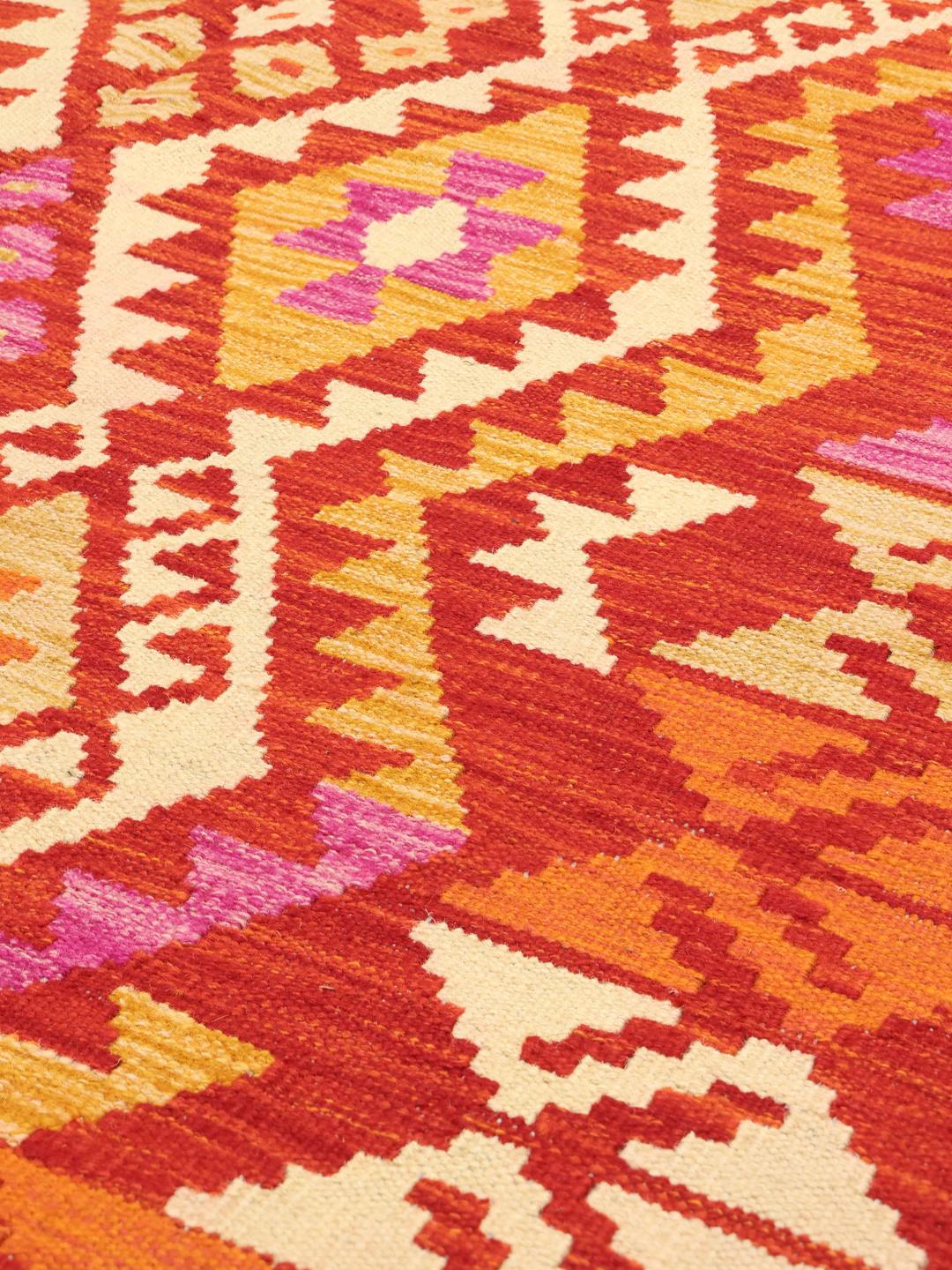A hand-woven, one-of-a-kind Afghan Kelim rug featuring a vibrant geometric pattern in red, orange, and pink tones.