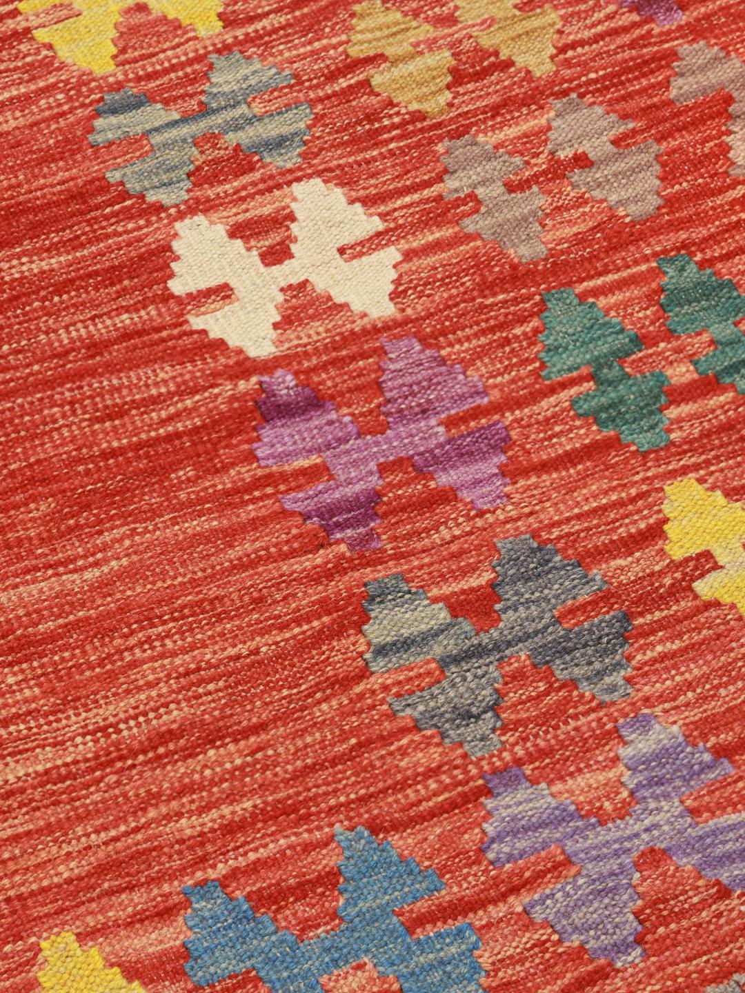 Een groot rechthoekig Afghaans Kelim-kleed met een levendig rood midden en een meerkleurige geometrische rand.